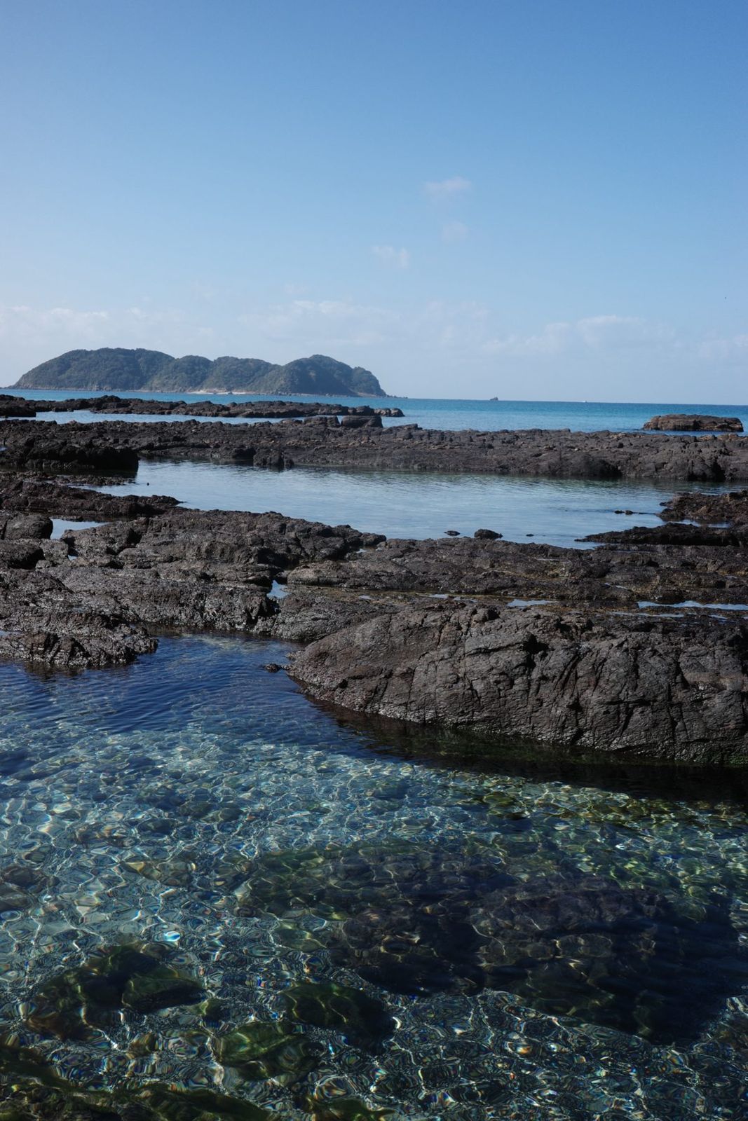 黒い岩場と透きとおった潮だまり、沖に浮かぶ島